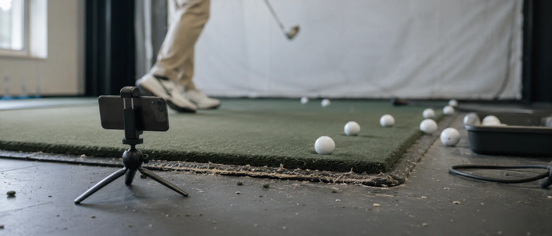 Phone on a low tripod beside an indoor hitting mat, the club head blurred mid-swing behind it